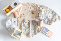 Flat lay of a SnuggleBox Infant (12-24 Months) collection featuring a unicorn-print top, floral shorts, a ruffled floral top, wooden play toys, and a polka-dot bib.