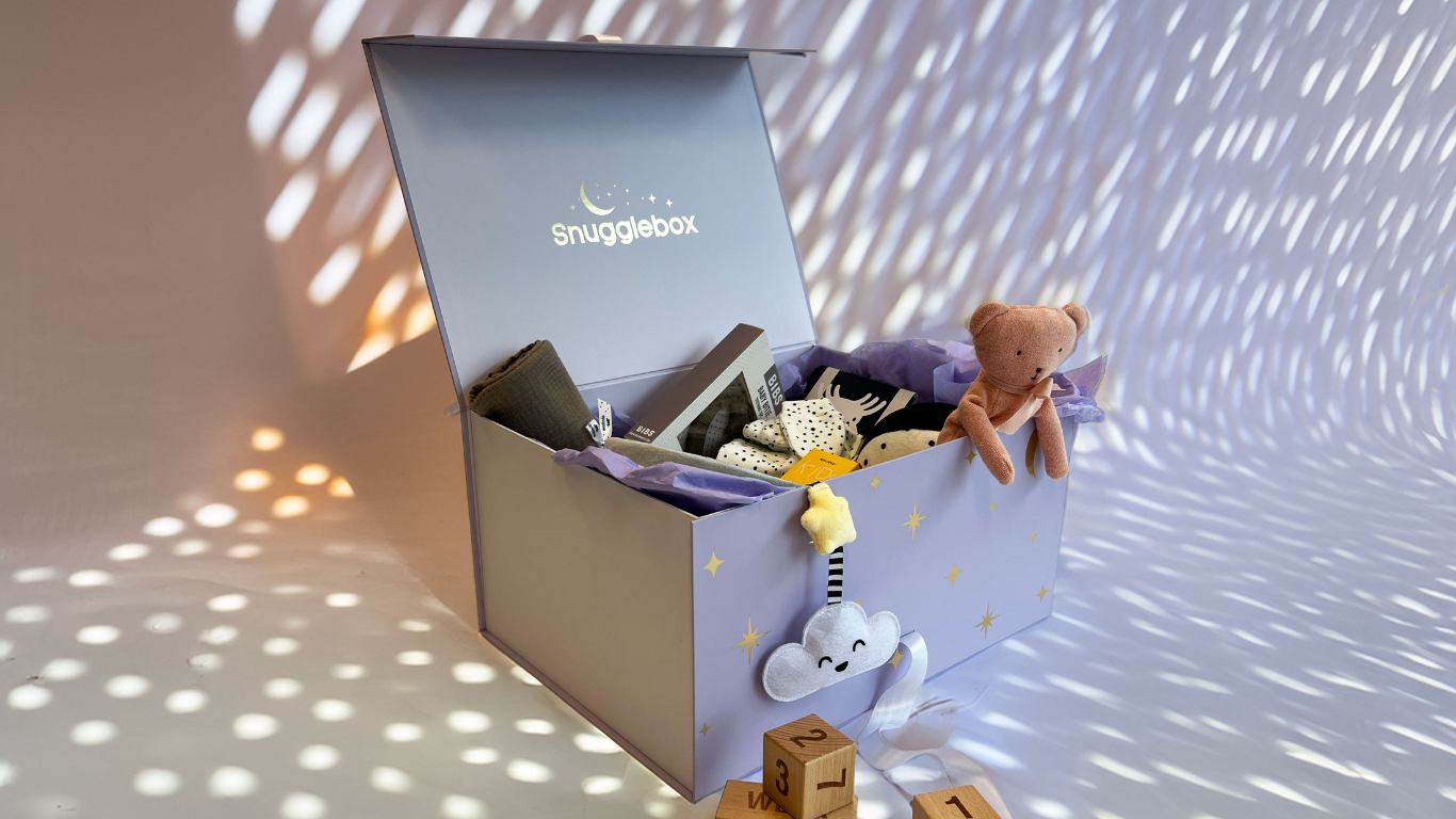 An open SnuggleBox filled with various baby products, including a plush toy, wooden blocks, and a cloud-shaped mobile, set against a magical purple background with soft lighting.